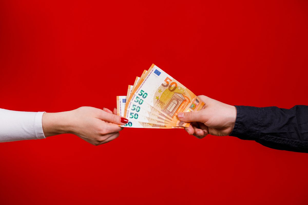 Braccia di due persone che si scambiano denaro (banconote da 50€) e sfondo rosso