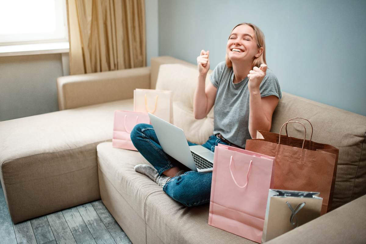 Ragazza felice dopo un acquisto online: seduta sul divano con pc e sacchetti shopping 